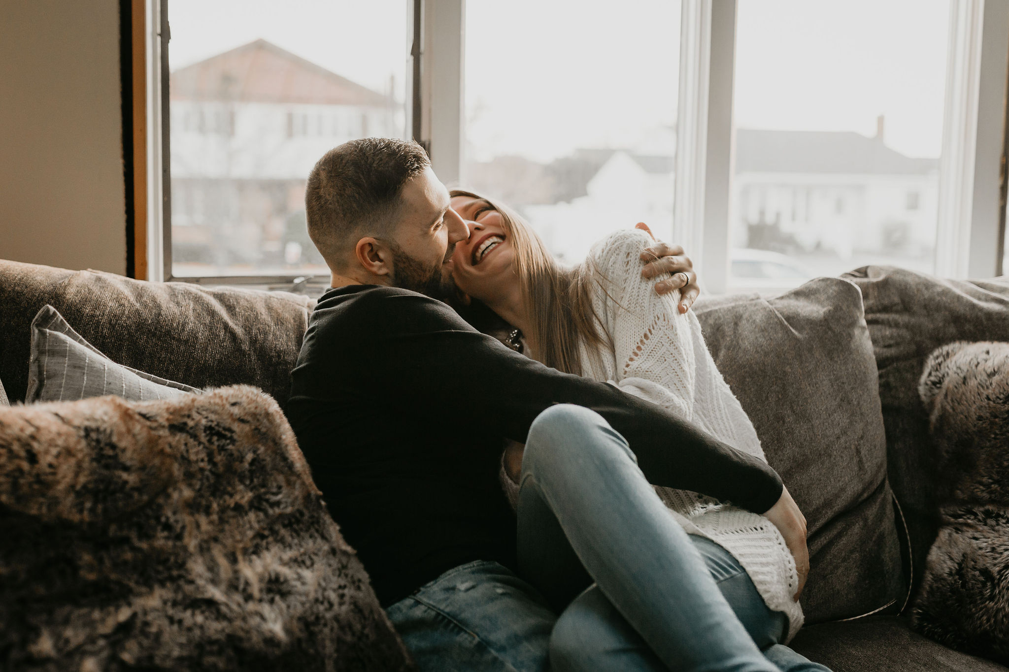 A couple snuggling on their couch smiling and laughing during their cozy in home lifestyle photo shoot on Long Island, New York.